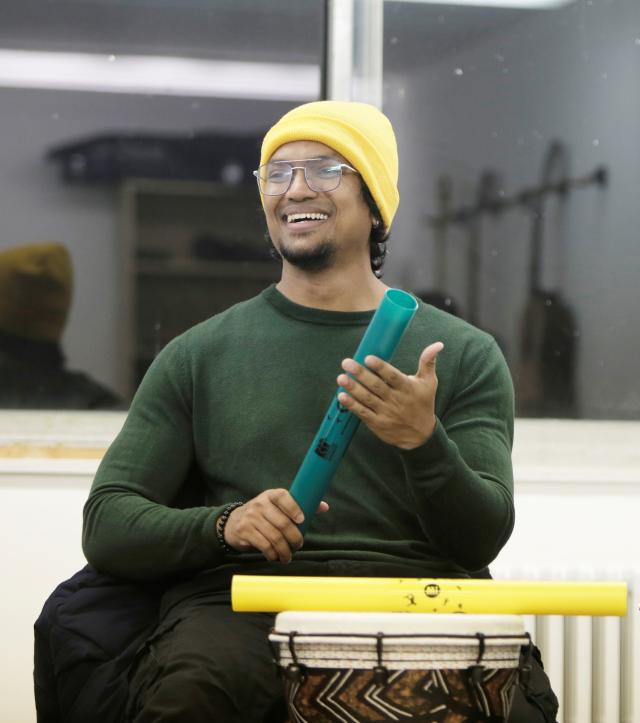 A seated, smiling man playing percussion instruments