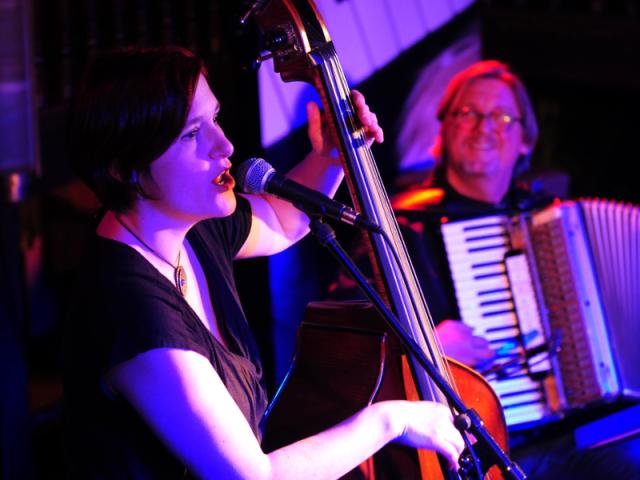 Une femme en premier plan qui chante accompagnée de sa contrebasse et un homme en second plan qui joue de l'accordéon