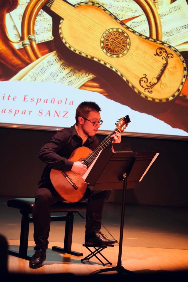 A seated man plays the guitar in front of a music stand